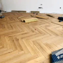 Montaż paneli podłogowych w jodełkę, widoczne deski, narzędzia i częściowo ułożona podłoga w jasnym odcieniu drewna, z pracownikiem w tle.