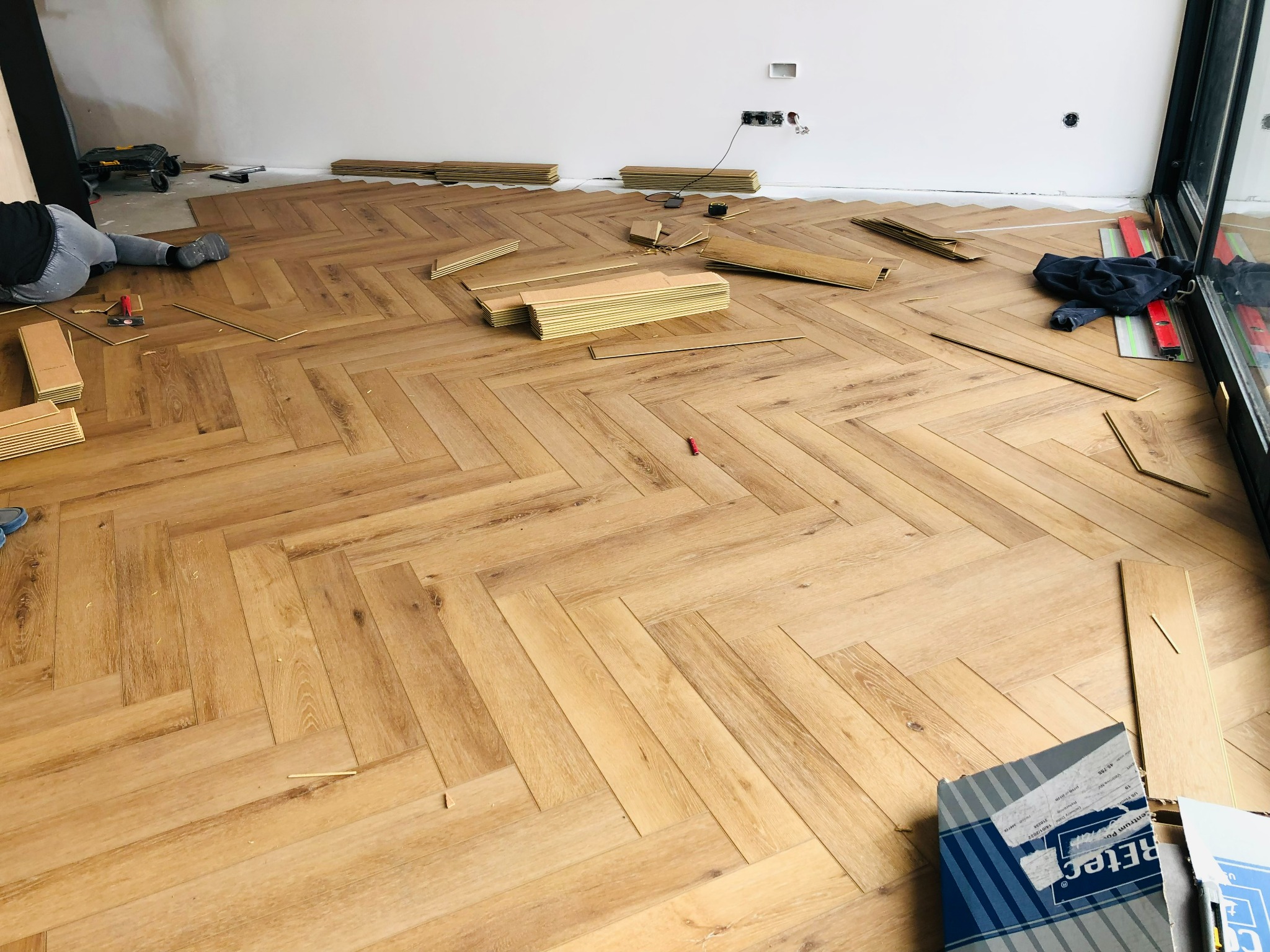 Montaż paneli podłogowych w jodełkę, widoczne deski, narzędzia i częściowo ułożona podłoga w jasnym odcieniu drewna, z pracownikiem w tle.