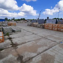 Składowisko płyt kamiennych i elementów nagrobnych w drewnianych skrzyniach na betonowym placu, widoczny dźwig budowlany w tle pod błękitnym niebem z obłokami.