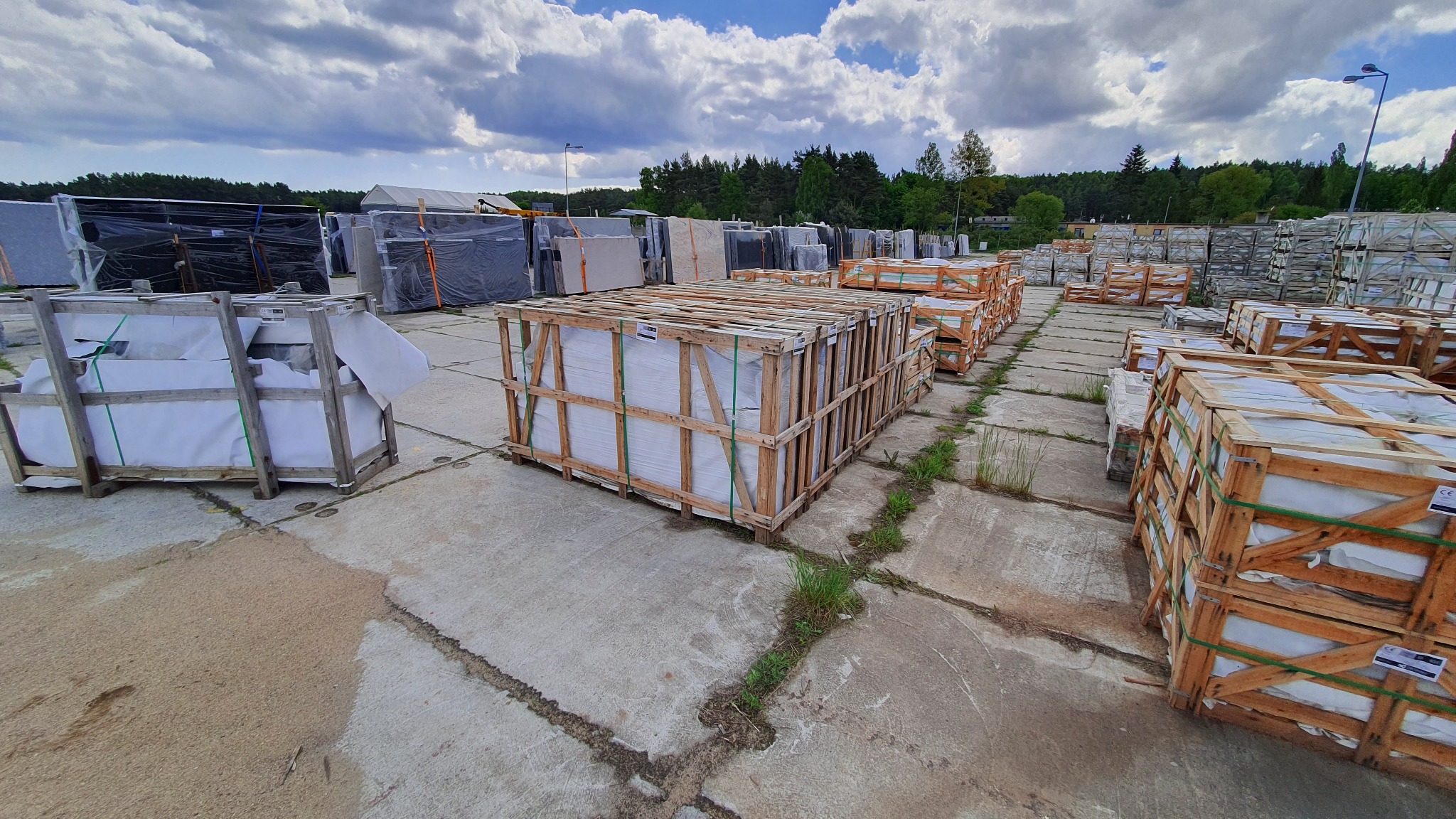 Materiały budowlane zapakowane w drewniane skrzynie, składowane na placu w Dębogórzu, widok na magazyn i zalesione tło pod pochmurnym niebem.