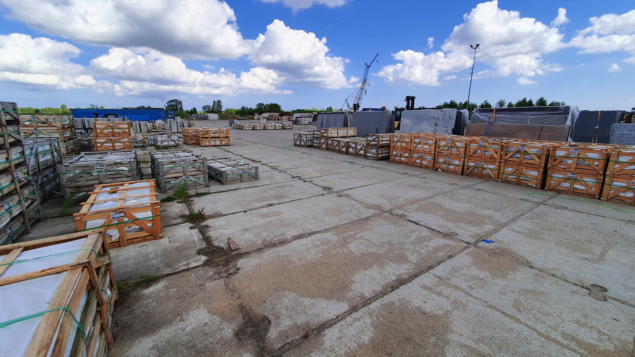 Składowisko płyt kamiennych i elementów nagrobnych w drewnianych skrzyniach na betonowym placu, widoczny dźwig budowlany w tle pod błękitnym niebem z obłokami.