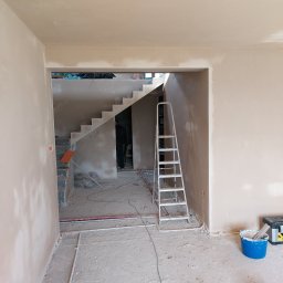 PERFEKTYNKI Patryk Jarząbski - Wnętrze w trakcie prac wykończeniowych, widoczne świeżo otynkowane ściany, schody w tle, drabina aluminiowa i narzędzia malarskie