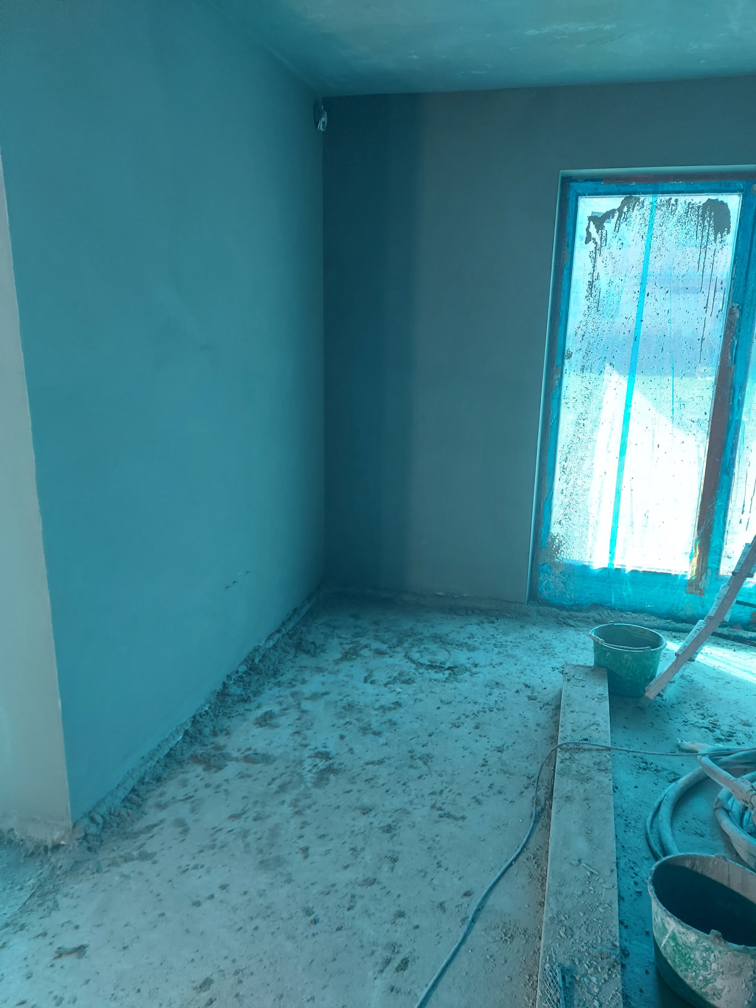 Świeżo otynkowane ściany w pomieszczeniu podczas prac wykończeniowych, okno zabezpieczone folią, widoczne narzędzia i wiaderka na podłodze.