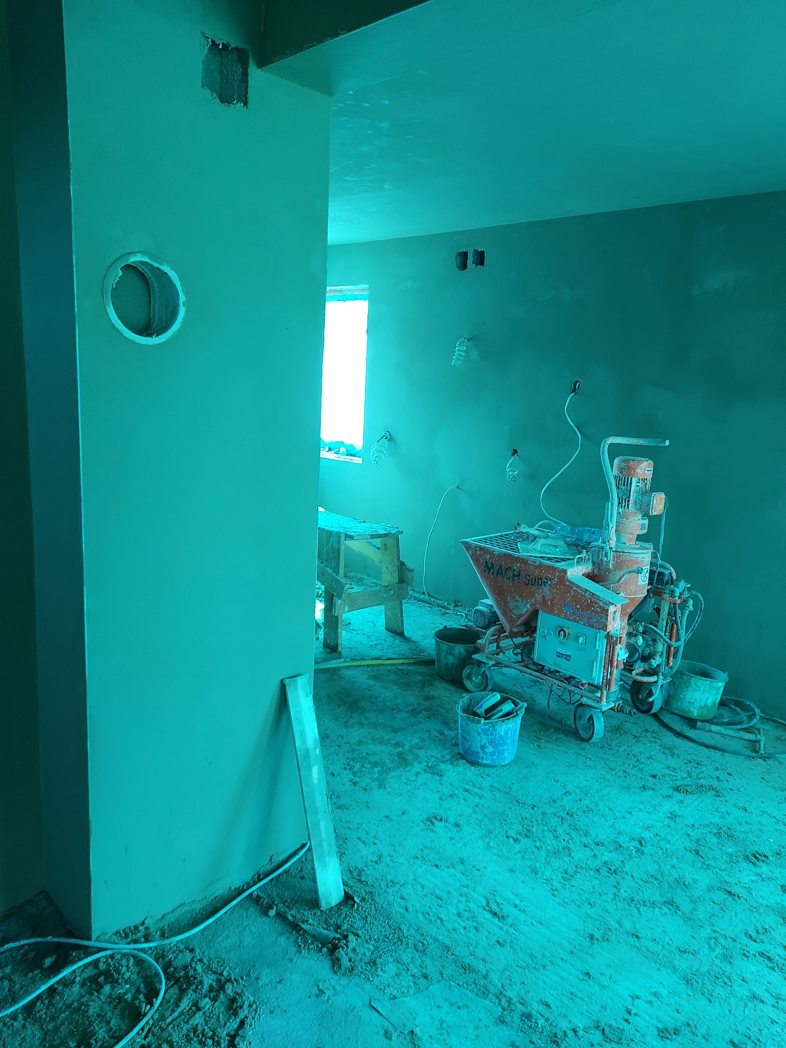 Wnętrze w trakcie prac wykończeniowych: ściany pokryte świeżym tynkiem, widoczna maszyna do tynkowania 'MACH Super' oraz narzędzia budowlane. Podłoga pokryta warstwą zaprawy.