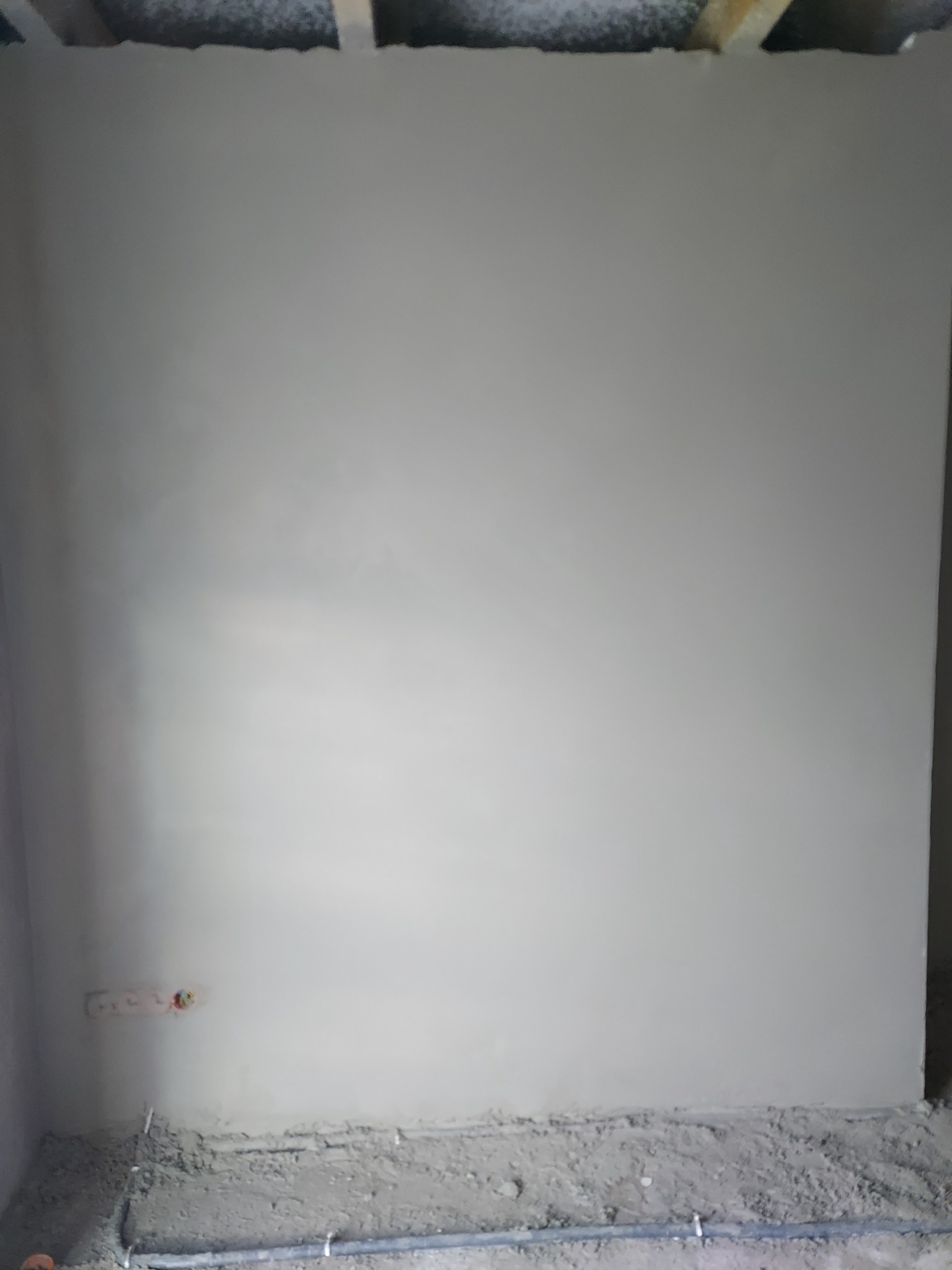 Świeżo nałożona, jeszcze wilgotna, jasnoszara warstwa na ścianie w trakcie prac wykończeniowych. Widoczne kable elektryczne wystające ze ściany, oraz rury przy podłodze.