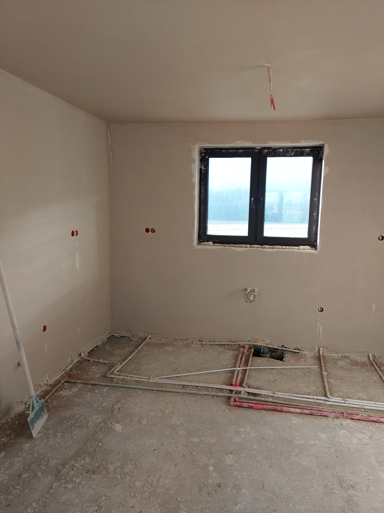 Pokój w trakcie prac wykończeniowych: ściany pokryte świeżym tynkiem, widoczne instalacje na podłodze, okno z czarną ramą, szpachelka oparta o ścianę.
