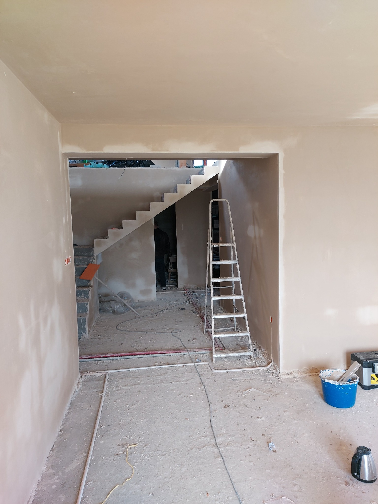 Wnętrze w trakcie prac wykończeniowych, widoczne świeżo otynkowane ściany, schody w tle, drabina aluminiowa i narzędzia malarskie