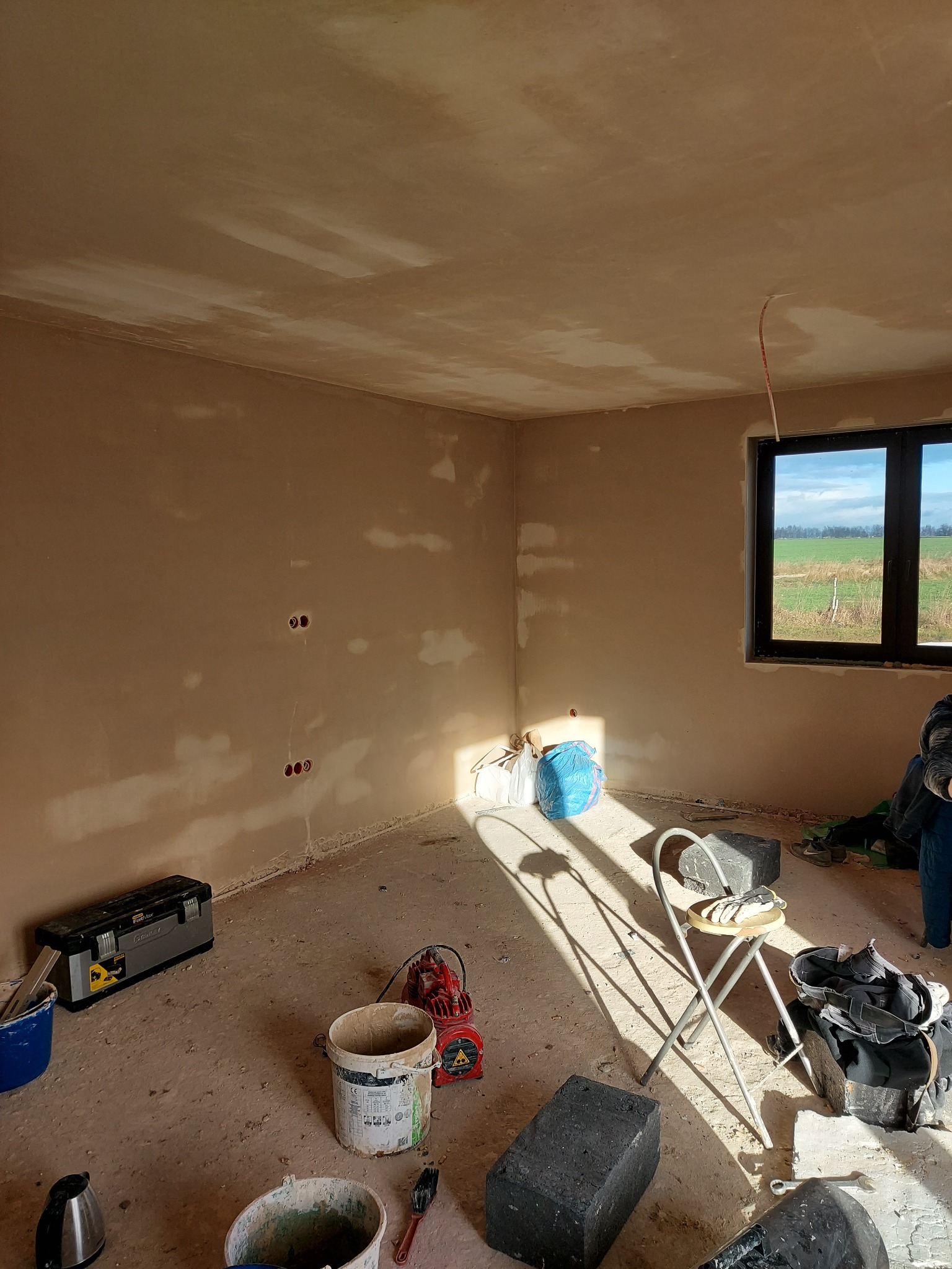 Wnętrze w trakcie prac w Białymstoku: ściany pokryte wstępną warstwą tynku, widoczne puszki elektryczne, narzędzia malarskie, drabina i okno z widokiem na pole.