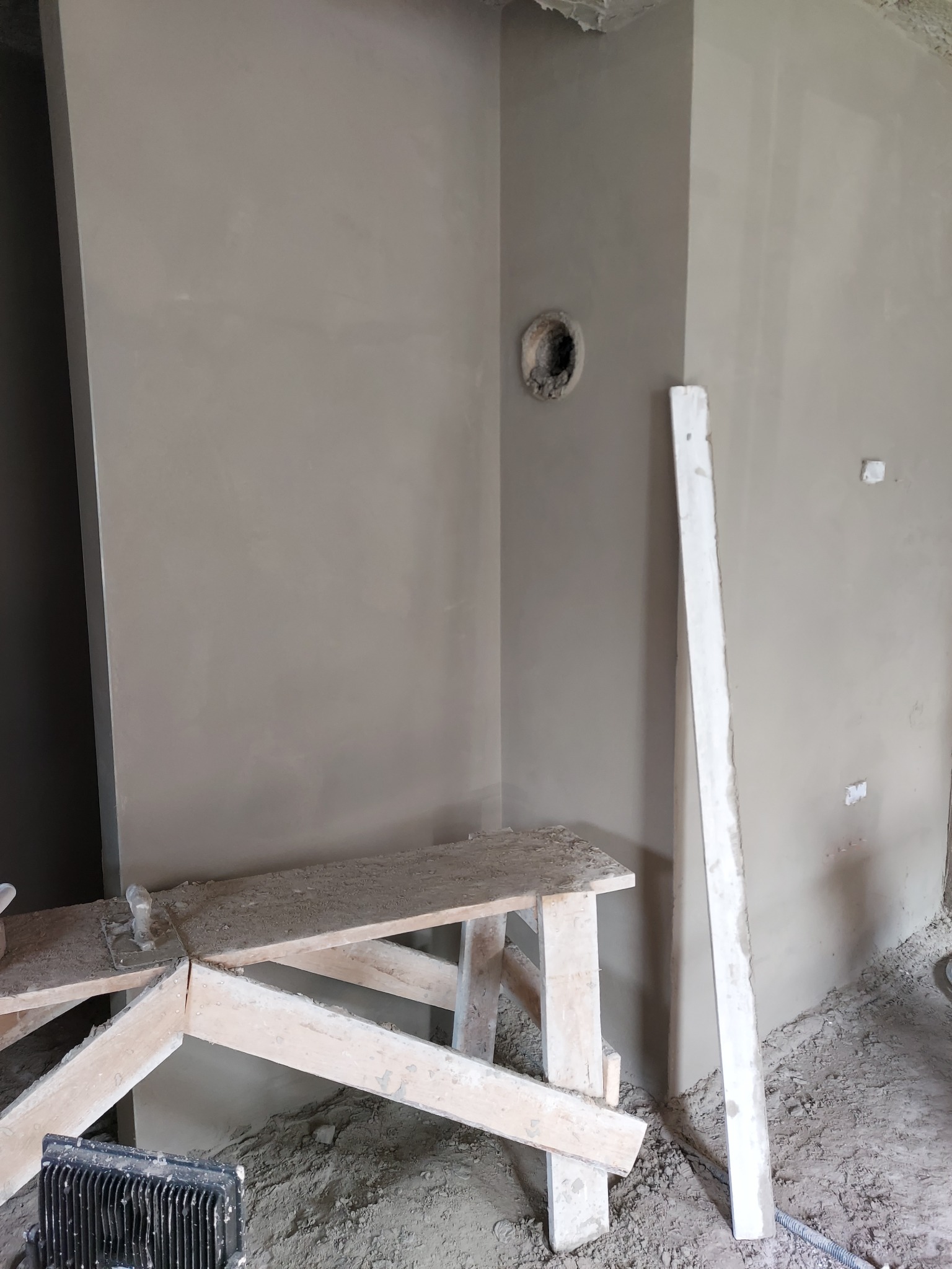 Świeżo nałożony tynk maszynowy na ścianach pomieszczenia w trakcie remontu, widoczny otwór pod puszkę elektryczną i drewniany kozioł roboczy pokryty resztkami zaprawy.
