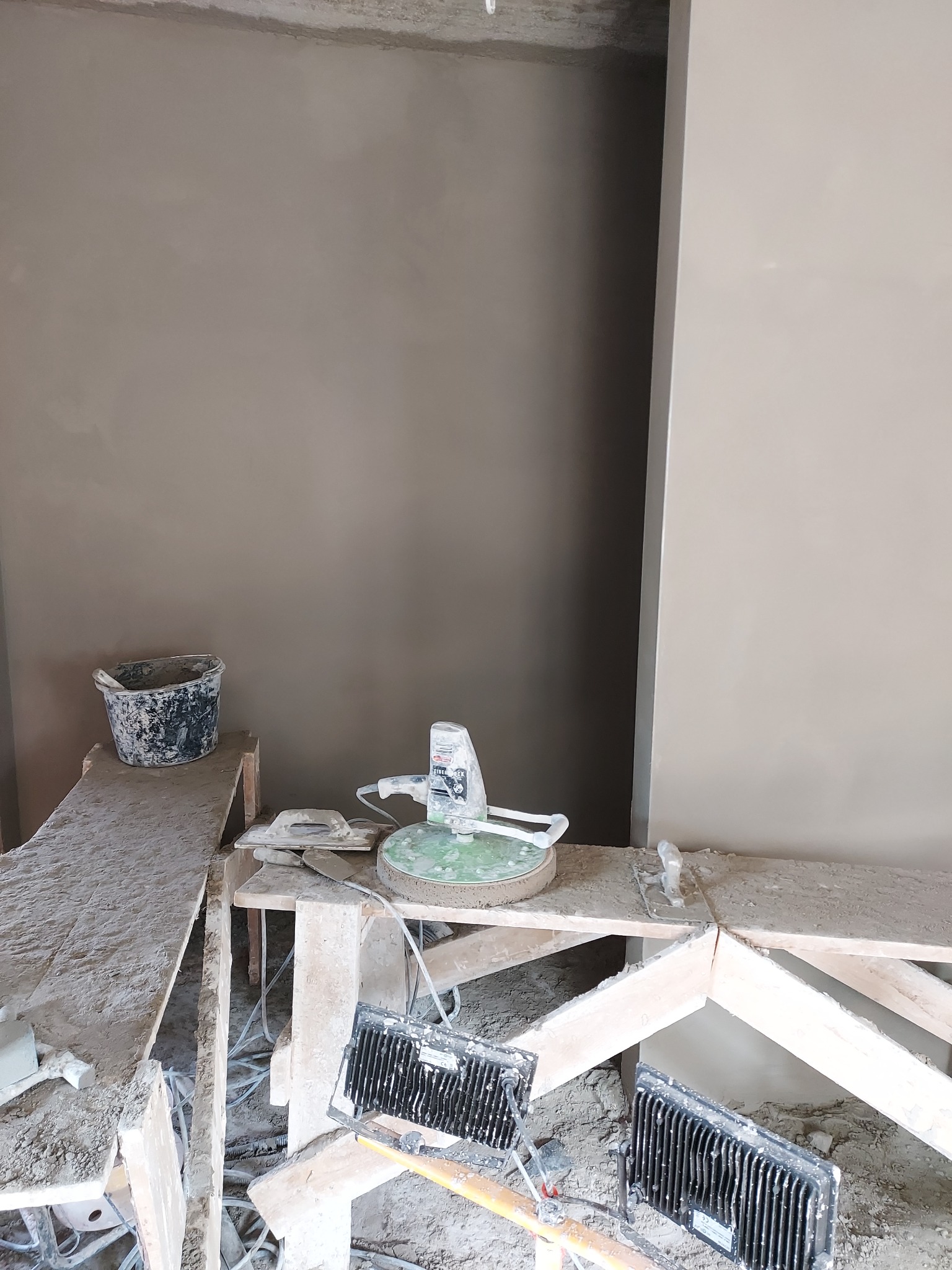 Świeżo otynkowana ściana w trakcie prac wykończeniowych, widoczna szlifierka do gładzi na drewnianym rusztowaniu, obok wiadro i szpachelki, dwa halogeny oświetlają miejsce pracy.