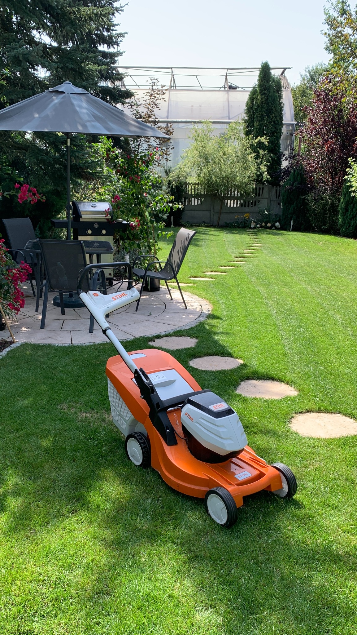 Pomarańczowa kosiarka akumulatorowa Stihl na trawniku, w tle grill, parasol, meble ogrodowe i ścieżka z kamieni prowadząca do szklarni.