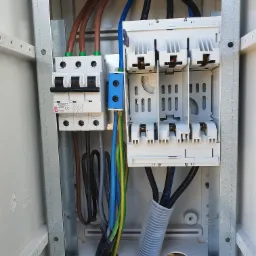 Otwarta skrzynka elektryczna z widocznymi bezpiecznikami, przewodami w różnych kolorach (brązowy, niebieski, czarny, zielono-żółty) oraz szarą rurą karbowaną, zamontowana wewnątrz obudowy...