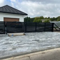 Nowoczesna posesja z przesuwną bramą wjazdową i poziomym, czarnym ogrodzeniem, wyłożona szarą kostką brukową, na tle domu z ciemnym dachem.