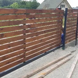 Nowoczesne ogrodzenie z poziomych desek w kolorze brązowym, na metalowych słupkach, z widocznymi elementami montażowymi i przygotowanymi deskami na ziemi.