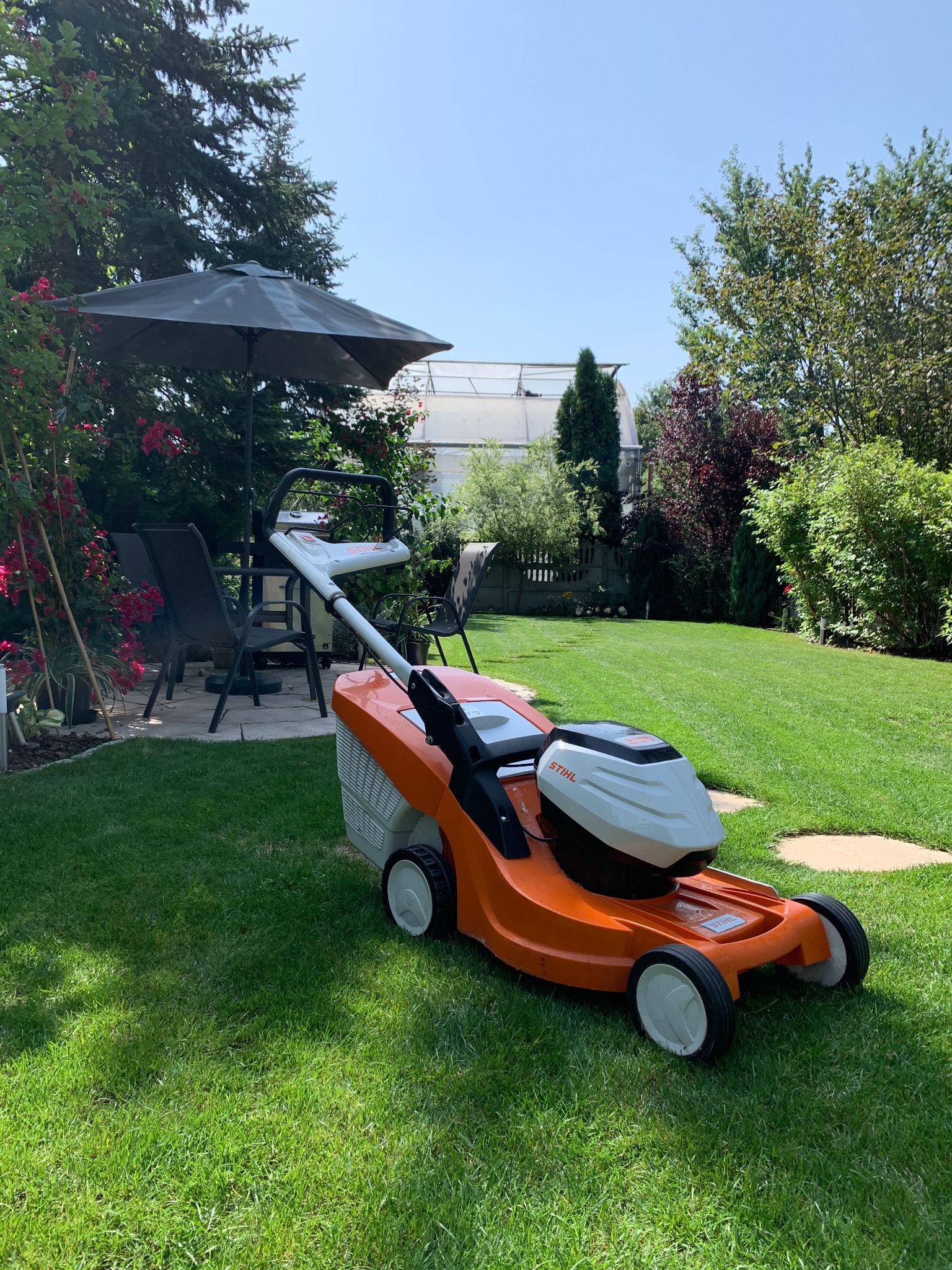 Pomarańczowa kosiarka marki Stihl na zadbanym, zielonym trawniku z widocznym zadaszeniem i roślinnością w tle.