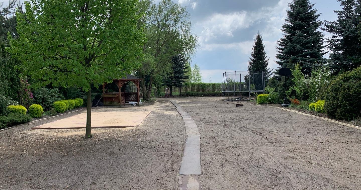 Przygotowany teren pod trawnik z wytyczoną ścieżką i miejscem na piaskownicę, otoczony zielenią.