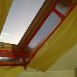 Okno dachowe w trakcie izolacji, zabezpieczone folią i taśmą, widoczne elementy konstrukcyjne poddasza.