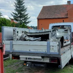 Uszkodzone elementy karoserii samochodu, przewożone na pace białego busa z otwartą skrzynią ładunkową, na tle zieleni i zabudowań.