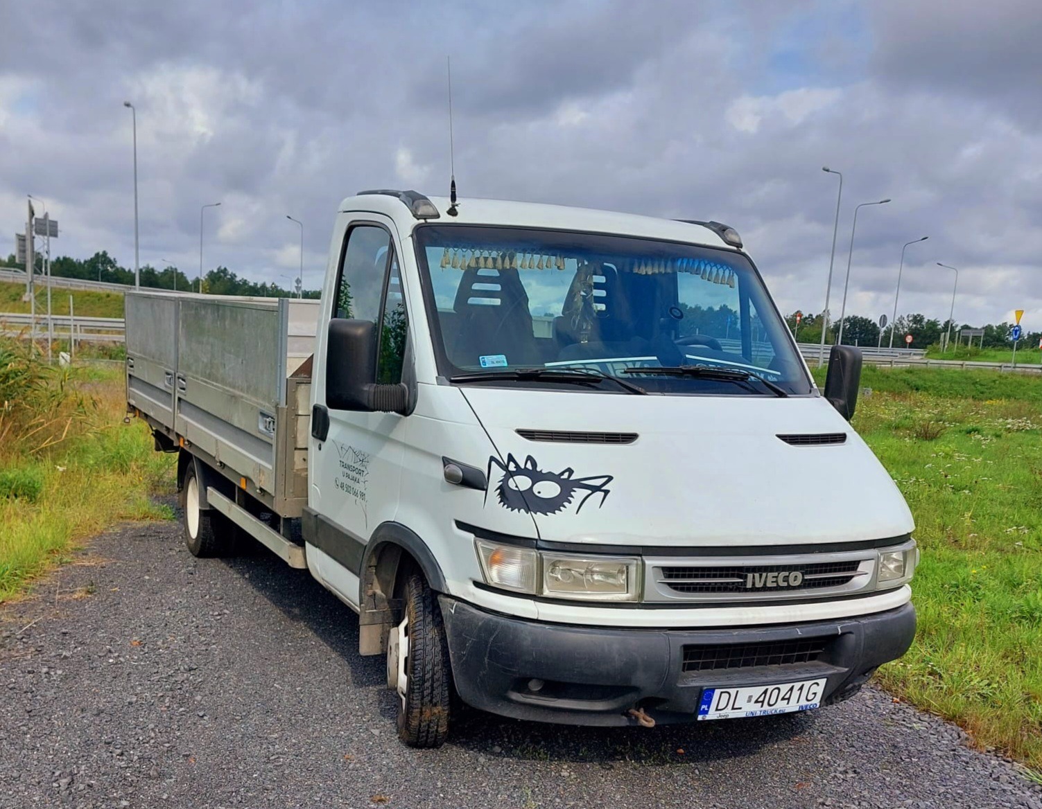 Biały samochód dostawczy Iveco z otwartą skrzynią ładunkową zaparkowany na żwirowej drodze, z czarnym rysunkiem pająka na masce, widoczne tablice rejestracyjne i numer telefonu firmy transportowej...