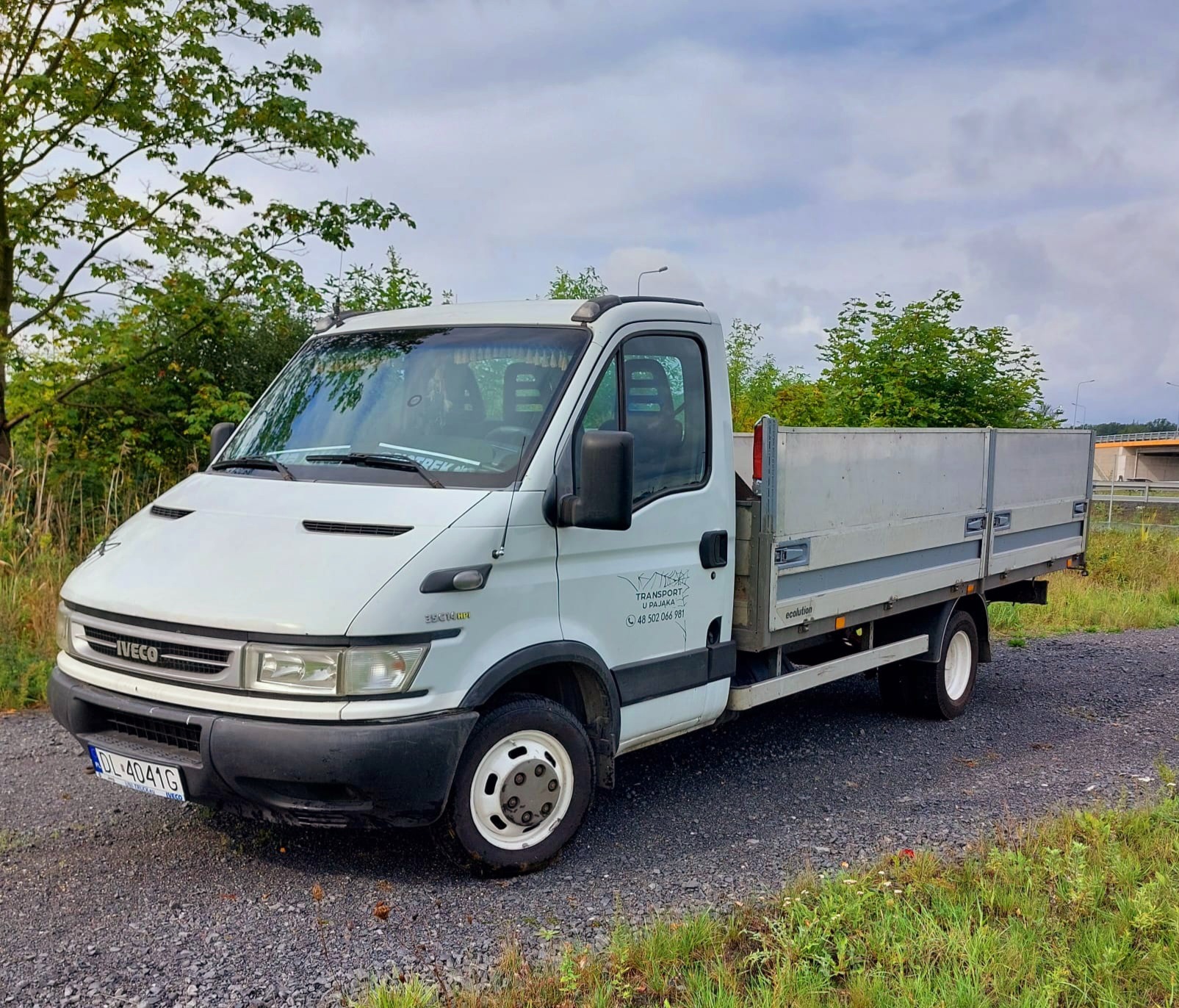 Biały samochód dostawczy Iveco z otwartą skrzynią ładunkową, zaparkowany na żwirowej drodze, z widocznym numerem rejestracyjnym i logo firmy transportowej na drzwiach.
