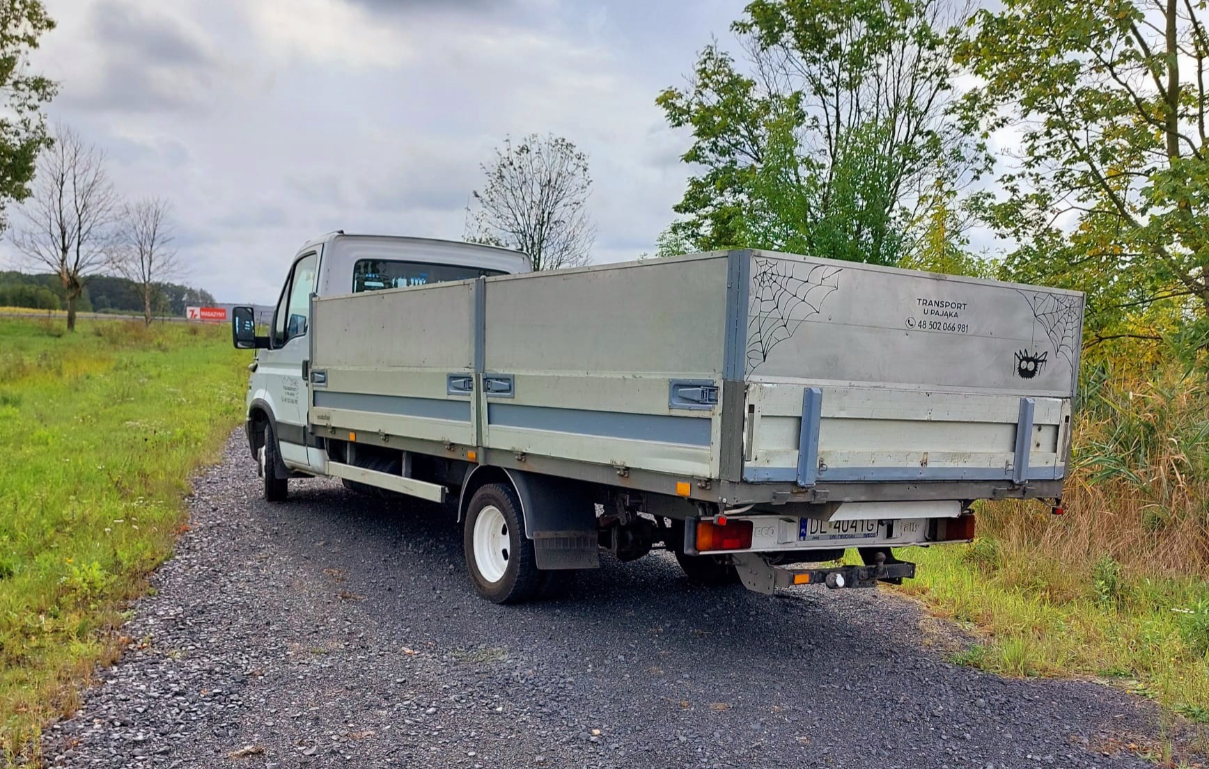 Tył białego samochodu dostawczego z napisem 'Transport u Pajaka' i rysunkami pajęczyn, stojącego na żwirowej drodze w otoczeniu zieleni.