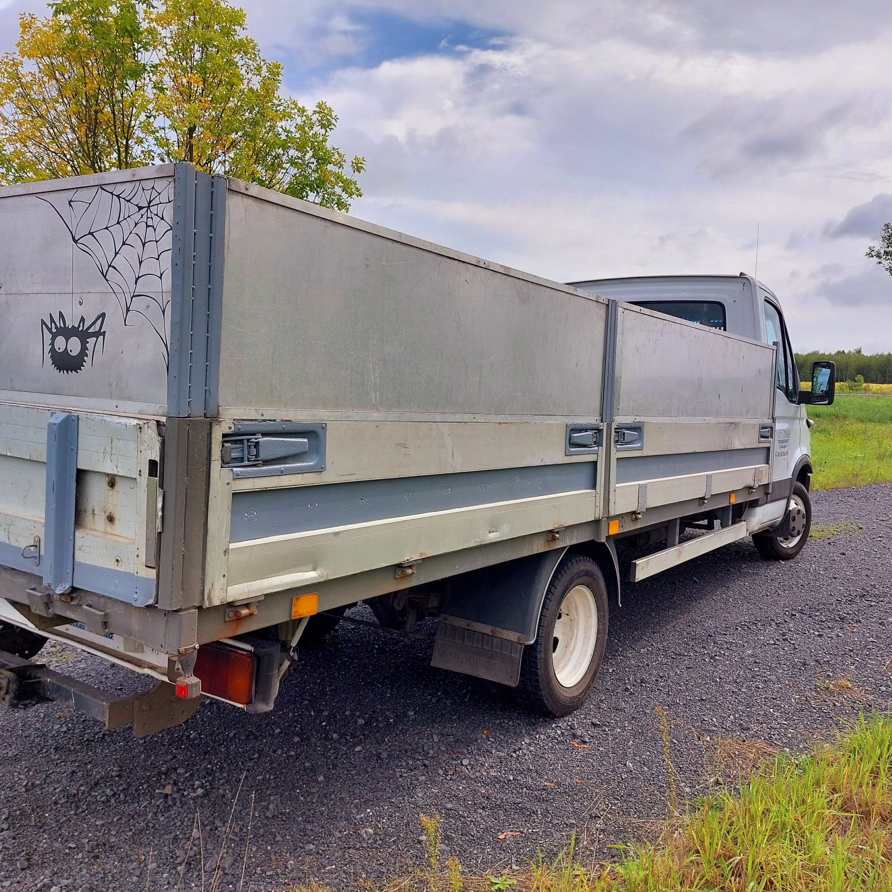 Szary samochód dostawczy z otwartą przestrzenią ładunkową, na której widnieje rysunek pająka w sieci, stojący na żwirowej drodze z widokiem na łąkę i drzewa.