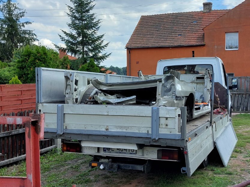 Uszkodzone elementy karoserii samochodu, przewożone na pace białego busa z otwartą skrzynią ładunkową, na tle zieleni i zabudowań.