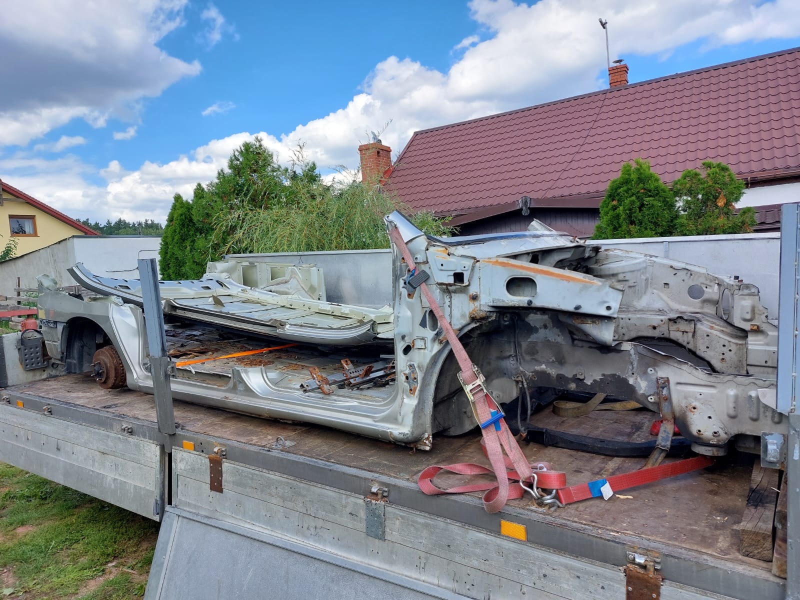Zdemontowany wrak samochodu osobowego, zabezpieczony pasami transportowymi na platformie lawety, widok z boku, w tle dom z czerwoną dachówką i zielona roślinność.