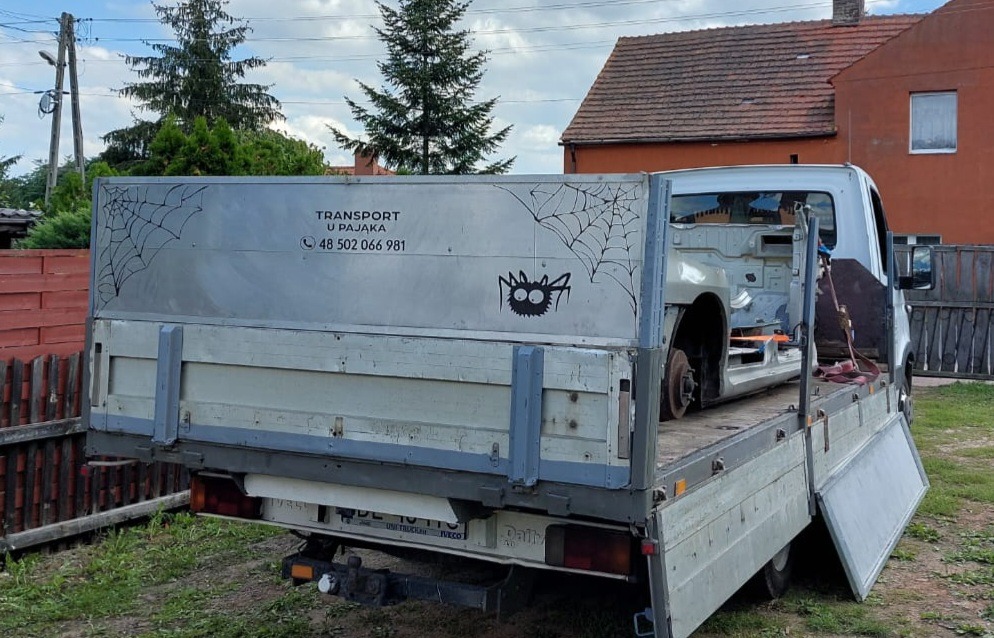 Otwarta skrzynia ładunkowa busa z naklejkami pająka i sieci, przewożąca karoserię innego pojazdu bez kół.
