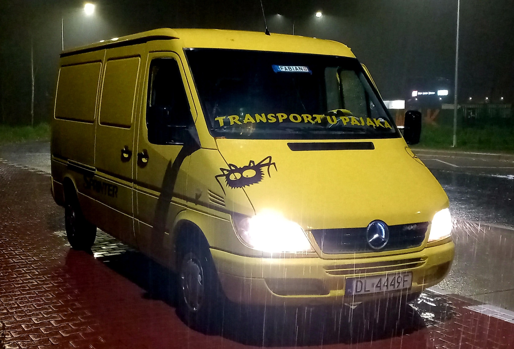 Żółty bus marki Mercedes Sprinter z napisem 'Transport u Pająka' na przedniej szybie i naklejką z wizerunkiem pająka na masce, sfotografowany nocą na mokrej nawierzchni.