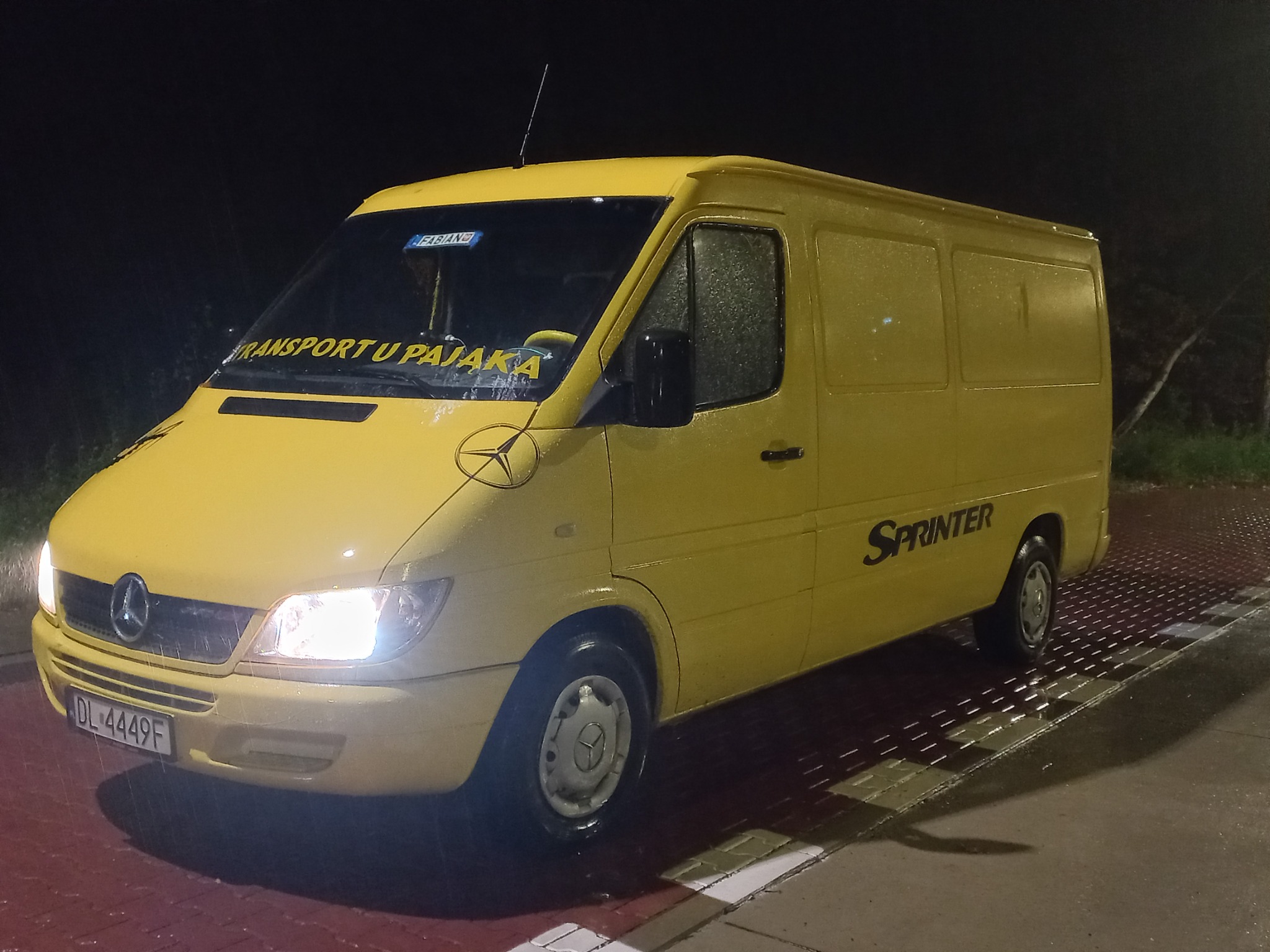 Żółty bus marki Mercedes Sprinter z napisem 'TRANSPORT U PAJĄKA' na przedniej szybie, zaparkowany na zewnątrz w nocy, widoczne krople deszczu na karoserii.