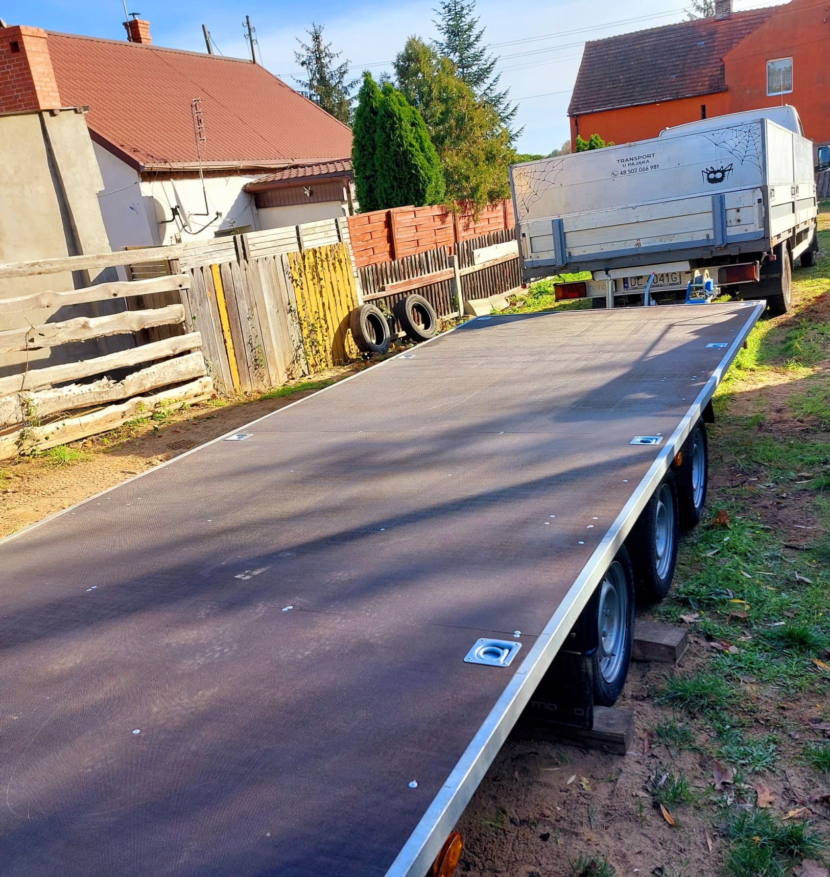 Pusta, czarna platforma lawety samochodowej z czterema kołami, zaparkowana na trawie z widocznym samochodem dostawczym w tle. Na skrzyni dostawczaka widoczny rysunek pajęczyny i kota.