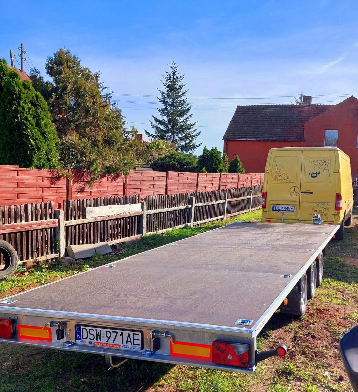 Płaska, trzyosiowa przyczepa samochodowa z żółtym busem marki Mercedes Sprinter w tle, zaparkowana na trawiastym terenie przed drewnianym płotem i czerwonym budynkiem.