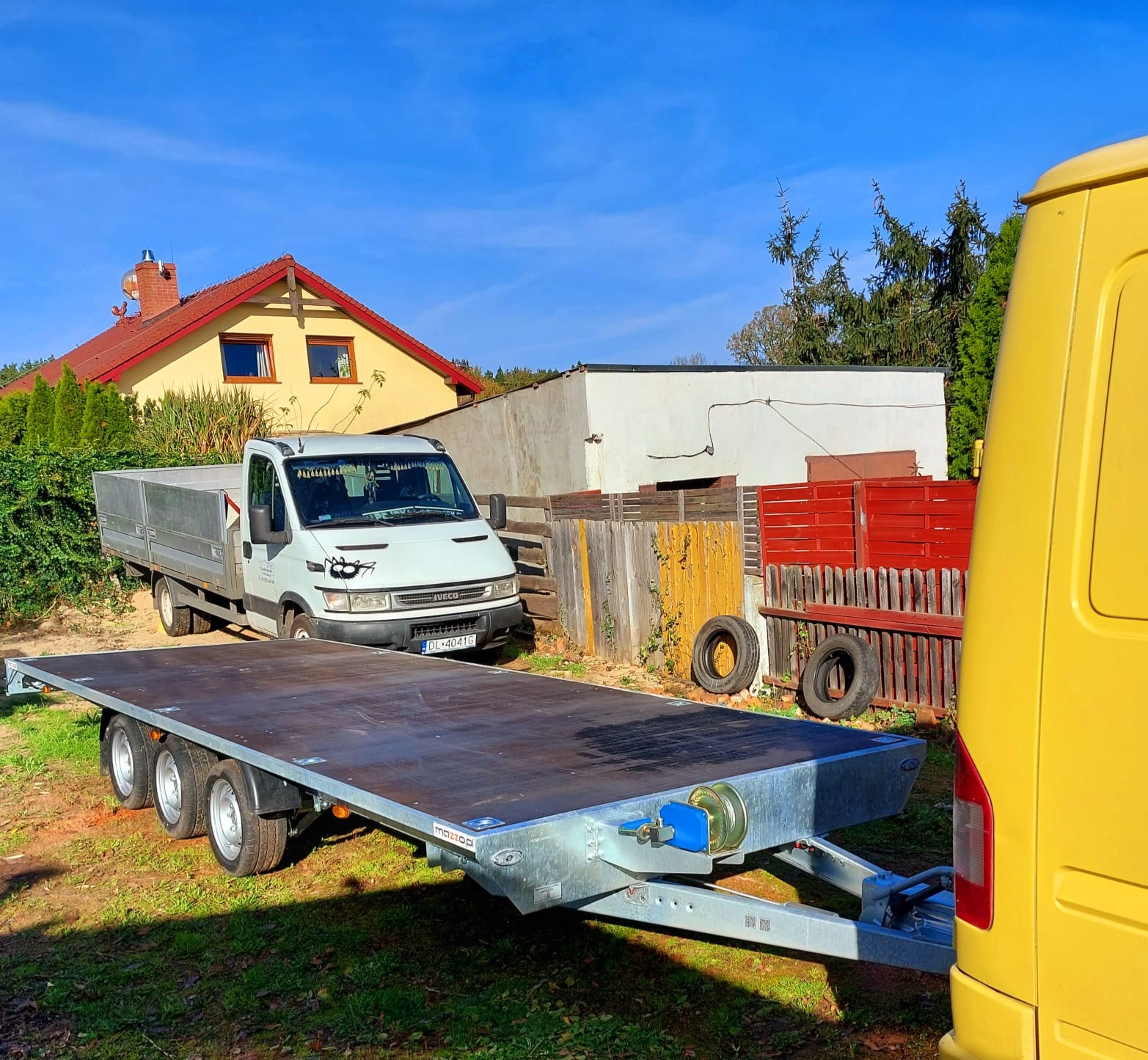 Płaska, trzyosiowa przyczepa transportowa z drewnianą podłogą, zaparkowana na trawie przed białym Iveco Daily z dekoracyjnymi naklejkami na reflektorach, na tle domu z czerwonym dachem i żółtą...