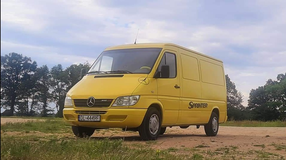 Żółty samochód dostawczy Mercedes-Benz Sprinter na polnej drodze, z widoczną tablicą rejestracyjną DL 4449F i napisem Sprinter na boku. W tle drzewa i pochmurne niebo.