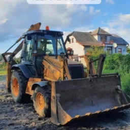 Żółta koparko-ładowarka CASE 580LE stoi na ubitej ziemi z widocznymi śladami opon, w tle dom jednorodzinny i błękitne niebo z chmurami. Na górze zdjęcia widnieje napis 'USŁUGI KOPARKO-ŁADOWARKĄ'...