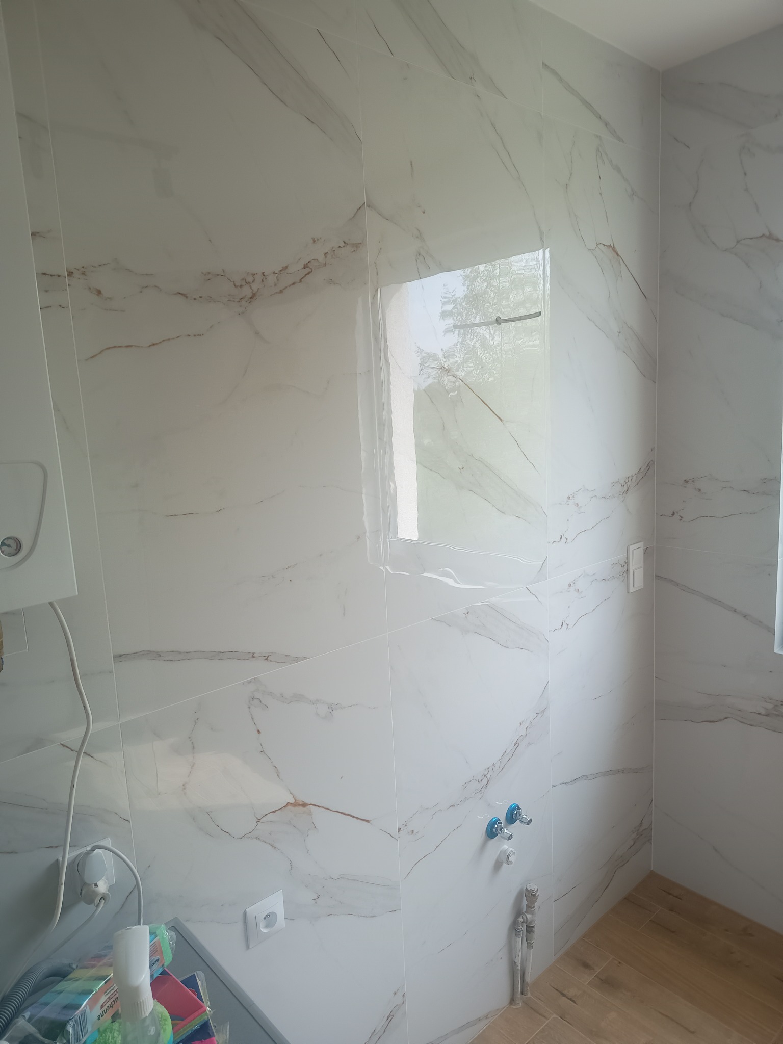 Wnętrze łazienki po remoncie: ściany wyłożone białymi płytkami imitującymi marmur z delikatnymi brązowymi żyłkami, widoczne przyłącza wodne z niebieskimi kurkami, fragment bojlera po lewej stronie...