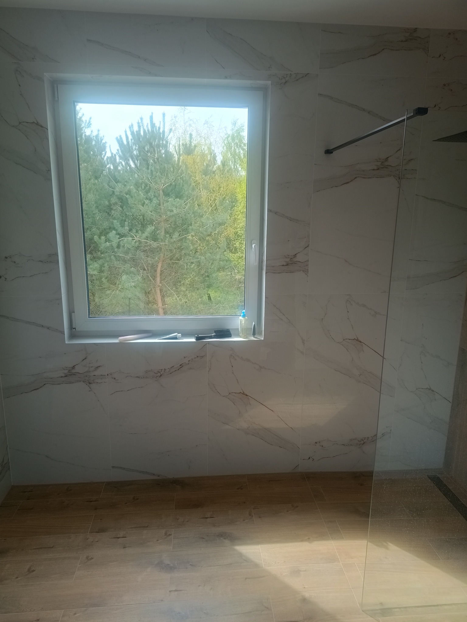 Wykończona łazienka z oknem, marmurowe płytki na ścianach, drewnopodobna podłoga, widok na las za oknem, minimalistyczny design kabiny prysznicowej.