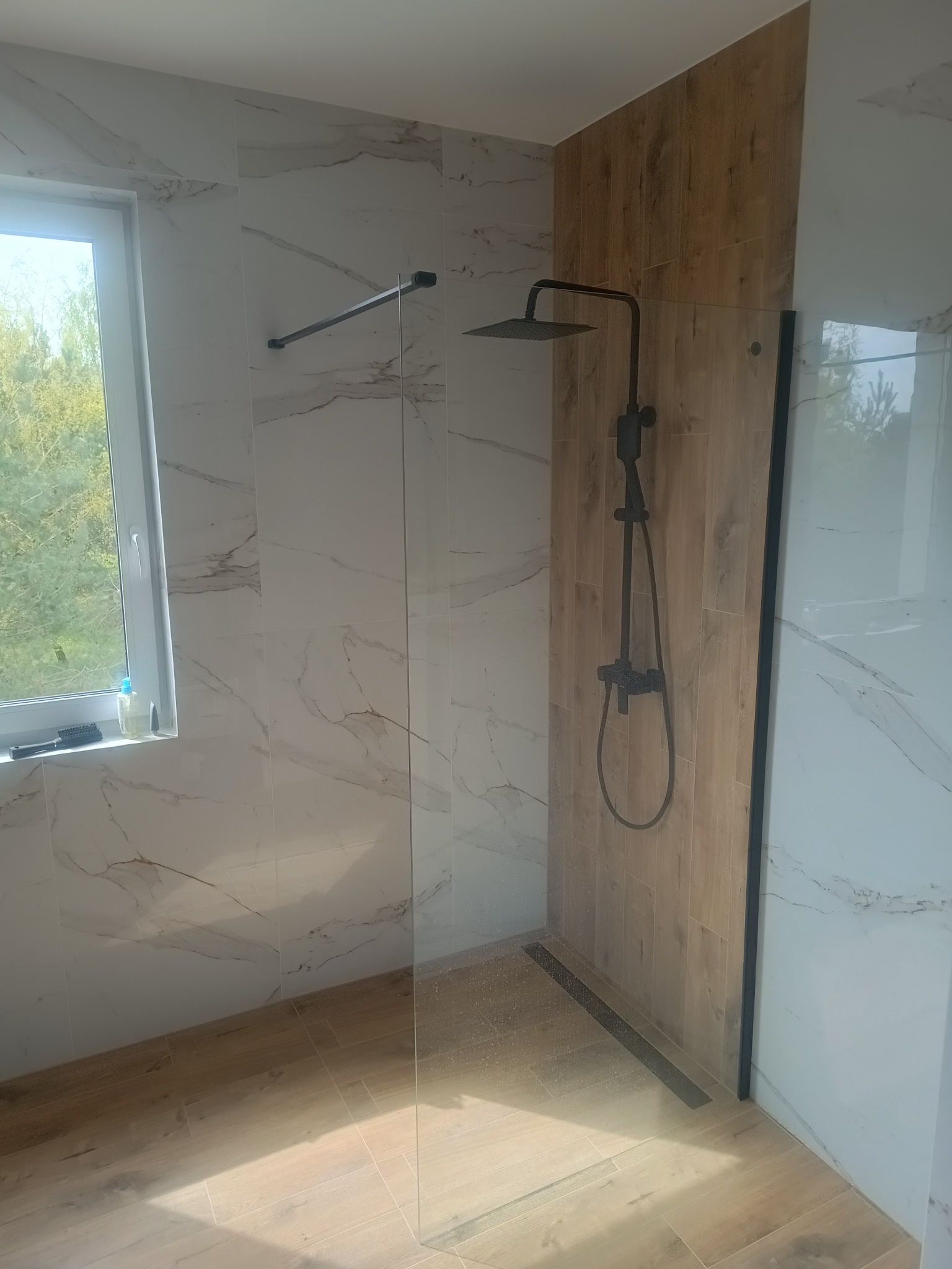 Nowoczesna kabina prysznicowa typu walk-in z czarną armaturą i odpływem liniowym, wykończona płytkami imitującymi marmur i drewno.