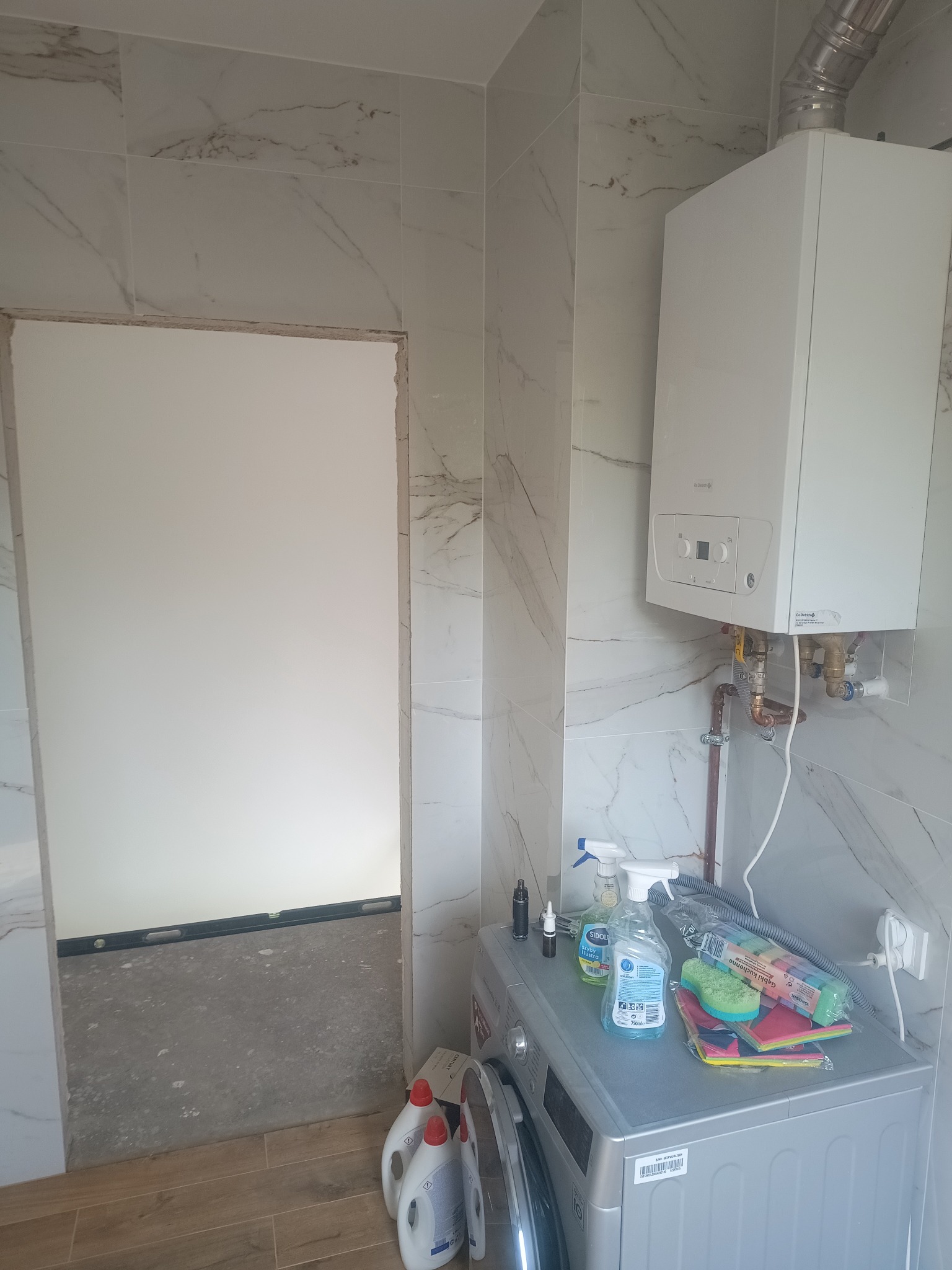 Łazienka w trakcie remontu: białe płytki imitujące marmur, piec gazowy na ścianie, pralka z środkami czystości i gąbkami na górze, widoczny fragment podłogi z paneli oraz przygotowane wejście...