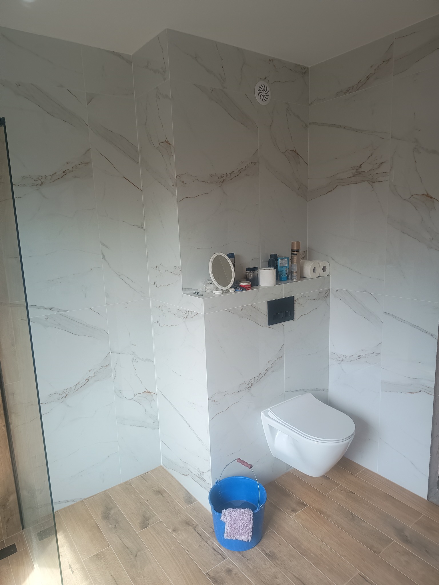 Nowoczesna łazienka z białymi płytkami imitującymi marmur, drewnopodobną podłogą i podwieszaną toaletą; na półce kosmetyki i lusterko, w rogu niebieskie wiadro z fioletową ścierką.