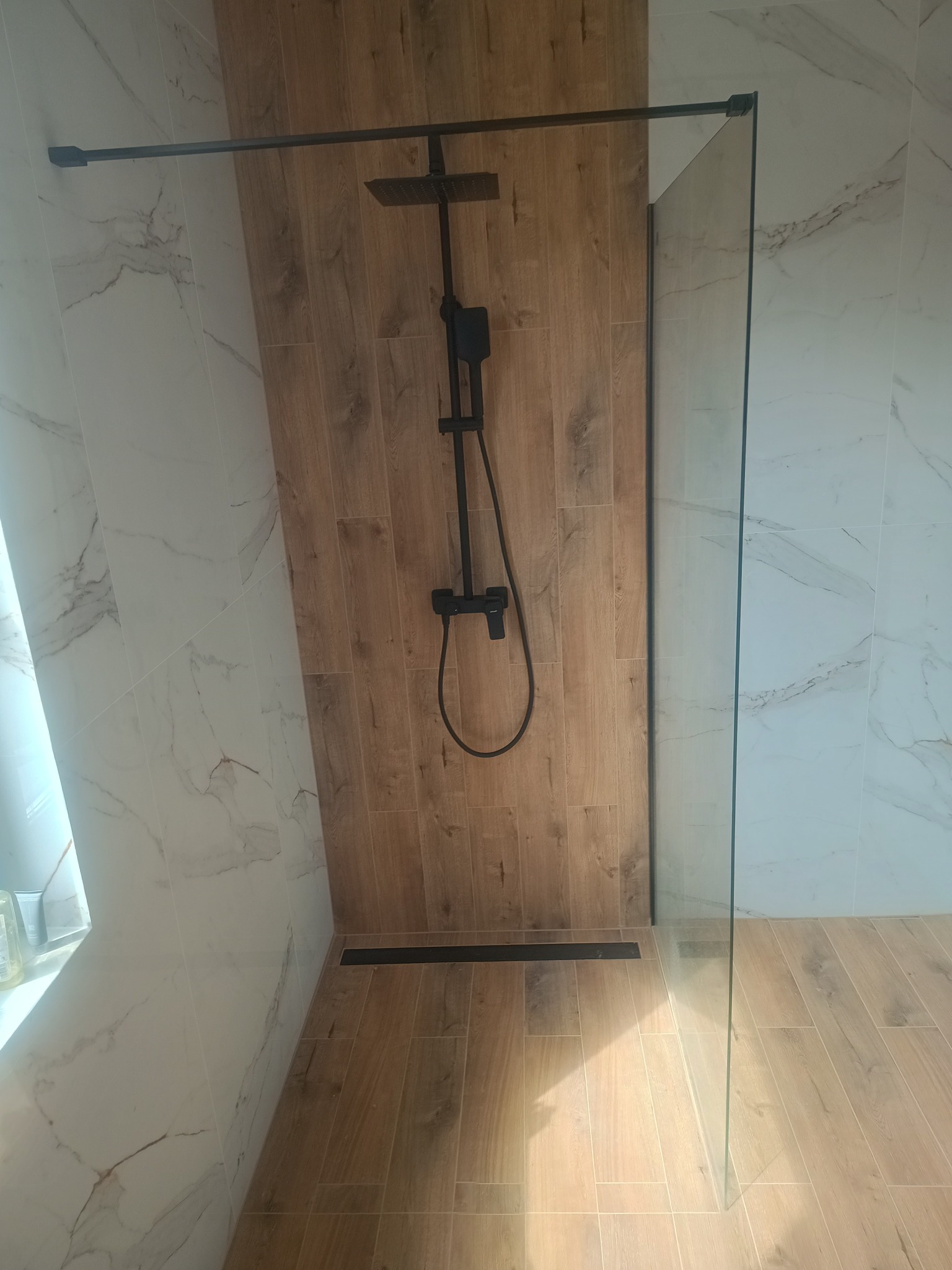 Nowoczesna kabina prysznicowa typu walk-in z czarną armaturą, drewnopodobnymi płytkami na ścianie i podłodze oraz białymi marmurkowymi płytkami na sąsiedniej ścianie.