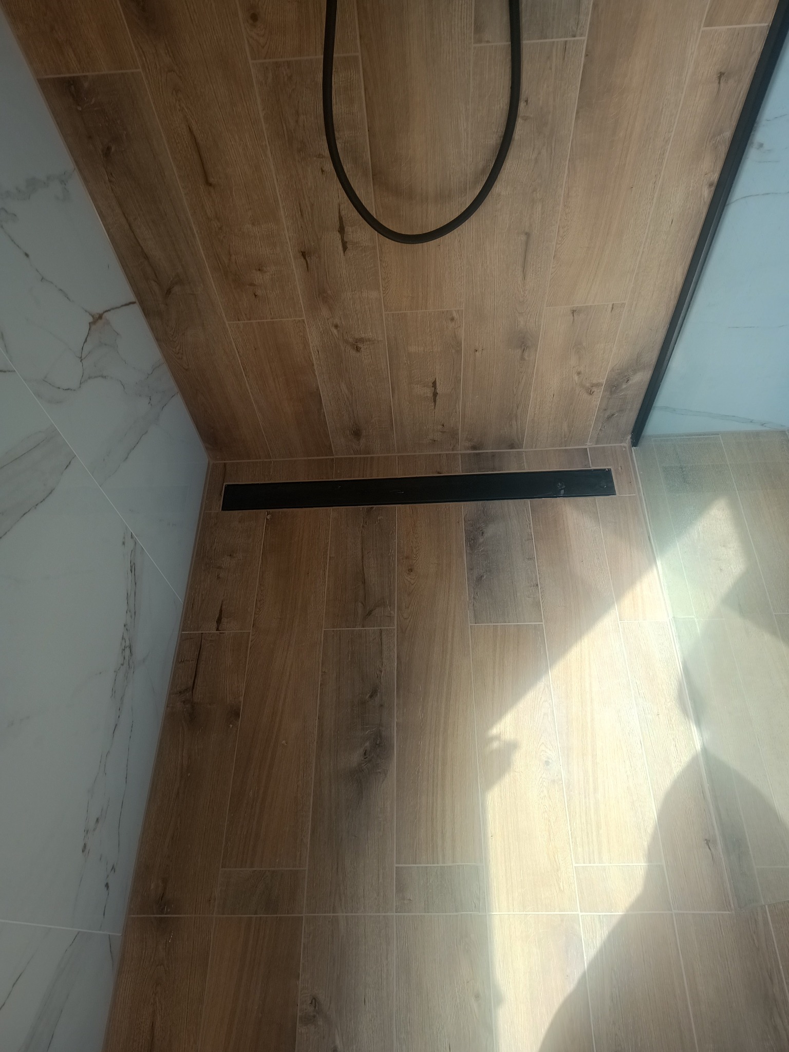 Wykończona podłoga w kabinie prysznicowej z drewnopodobnych płytek ceramicznych, z czarnym odpływem liniowym i fragmentem węża prysznicowego w górnej części kadru.