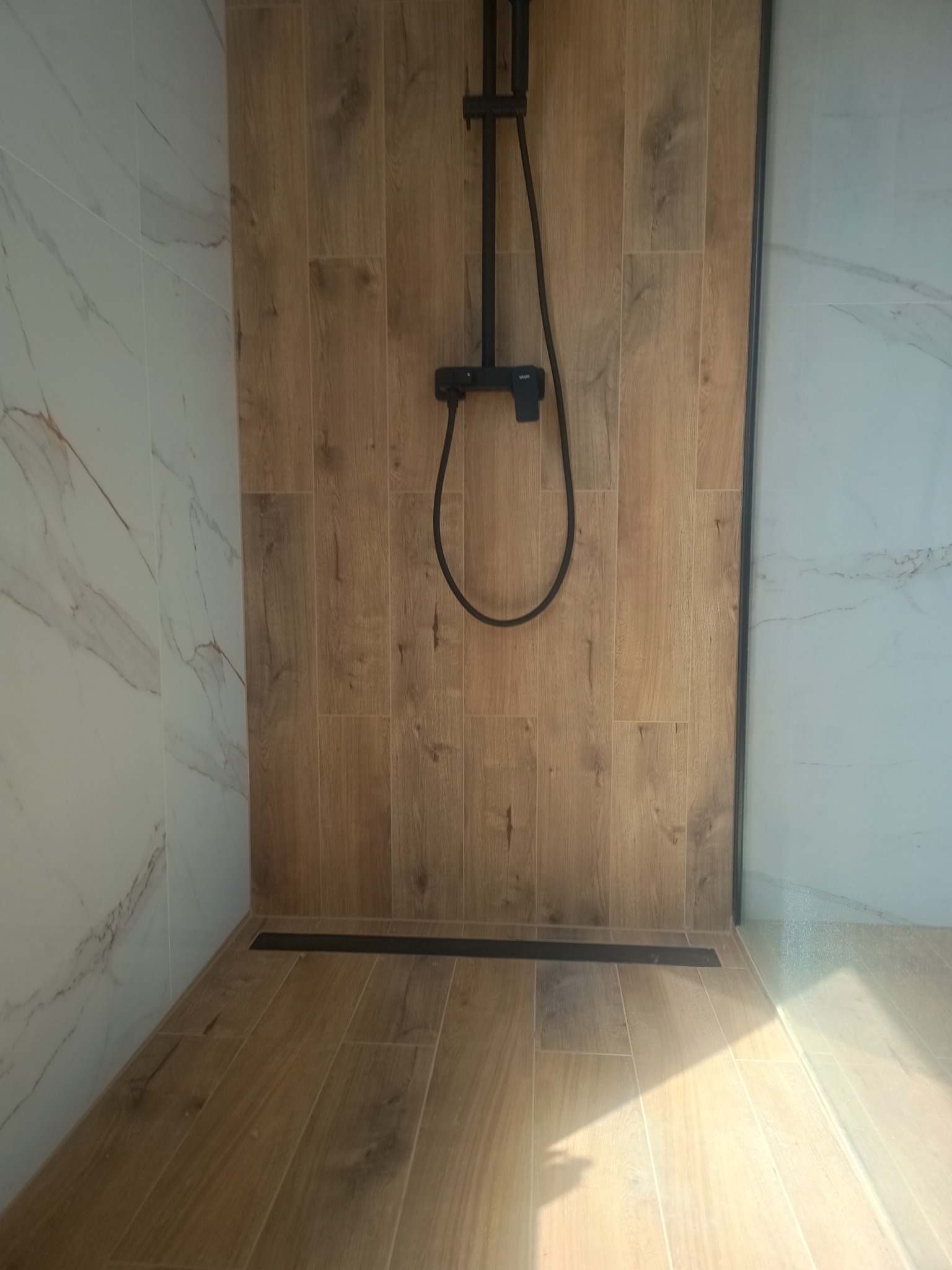 Nowoczesna kabina prysznicowa z armaturą w kolorze czarnym, wyłożona płytkami imitującymi drewno i marmur, z odpływem liniowym i delikatnym oświetleniem.