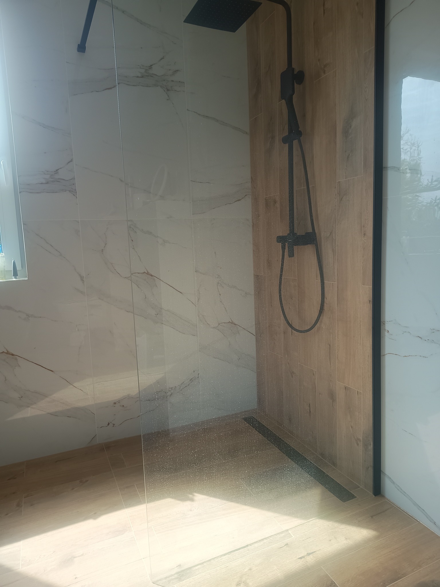 Nowoczesna kabina prysznicowa walk-in z czarną armaturą, wykończona płytkami imitującymi marmur i drewno, z widocznym odpływem liniowym.