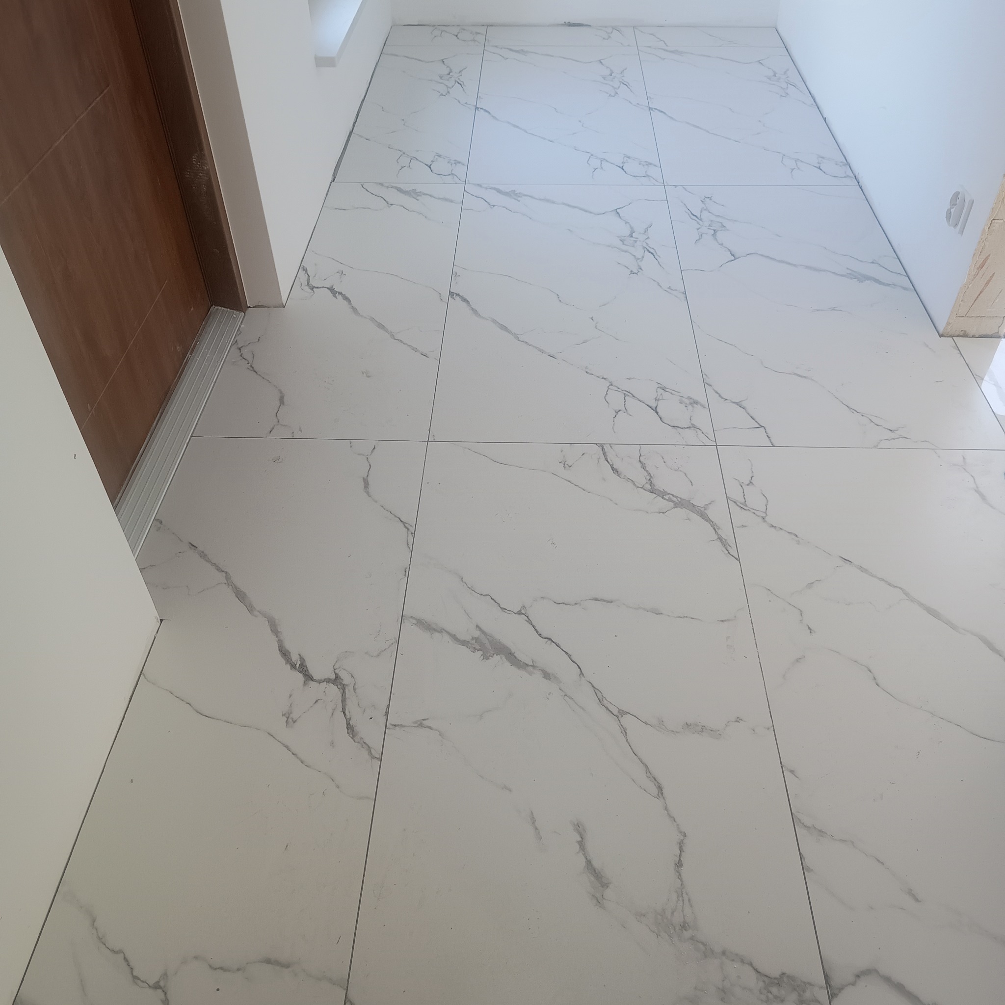 Białe płytki podłogowe z szarym marmurkowym wzorem ułożone w nowym pomieszczeniu, widoczna część brązowych drzwi z aluminiową listwą progową.