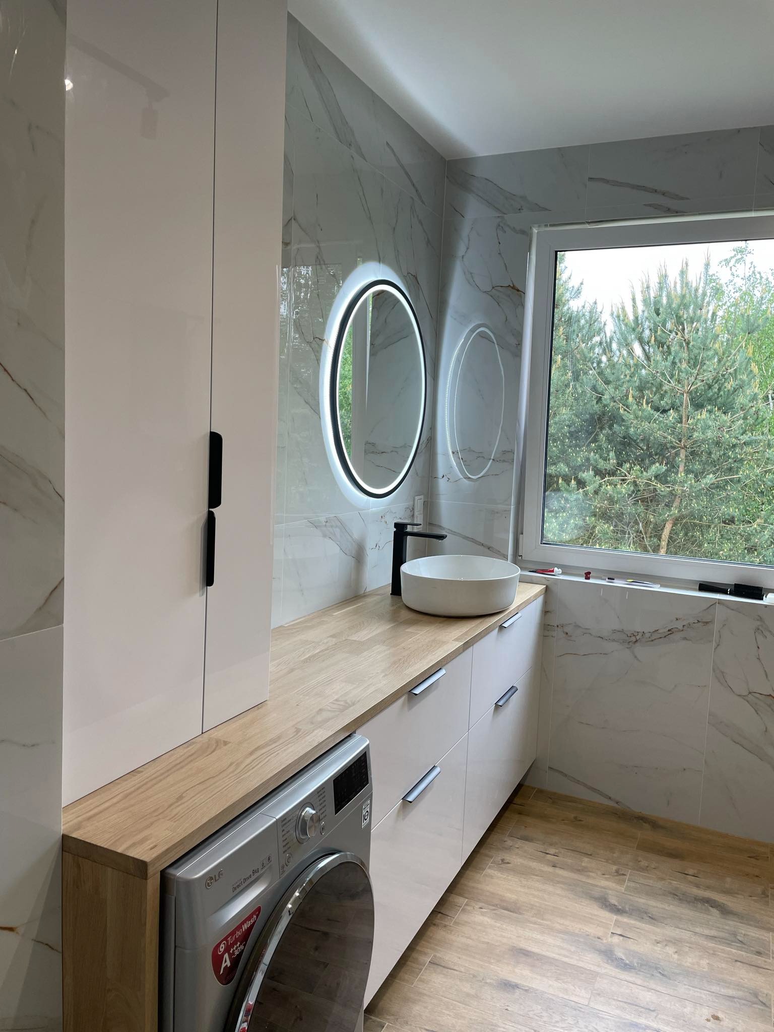 Nowoczesna łazienka z marmurowymi płytkami, okrągłym lustrem z podświetleniem LED, drewnianym blatem, białą umywalką nablatową, pralką LG i drewnopodobną podłogą.