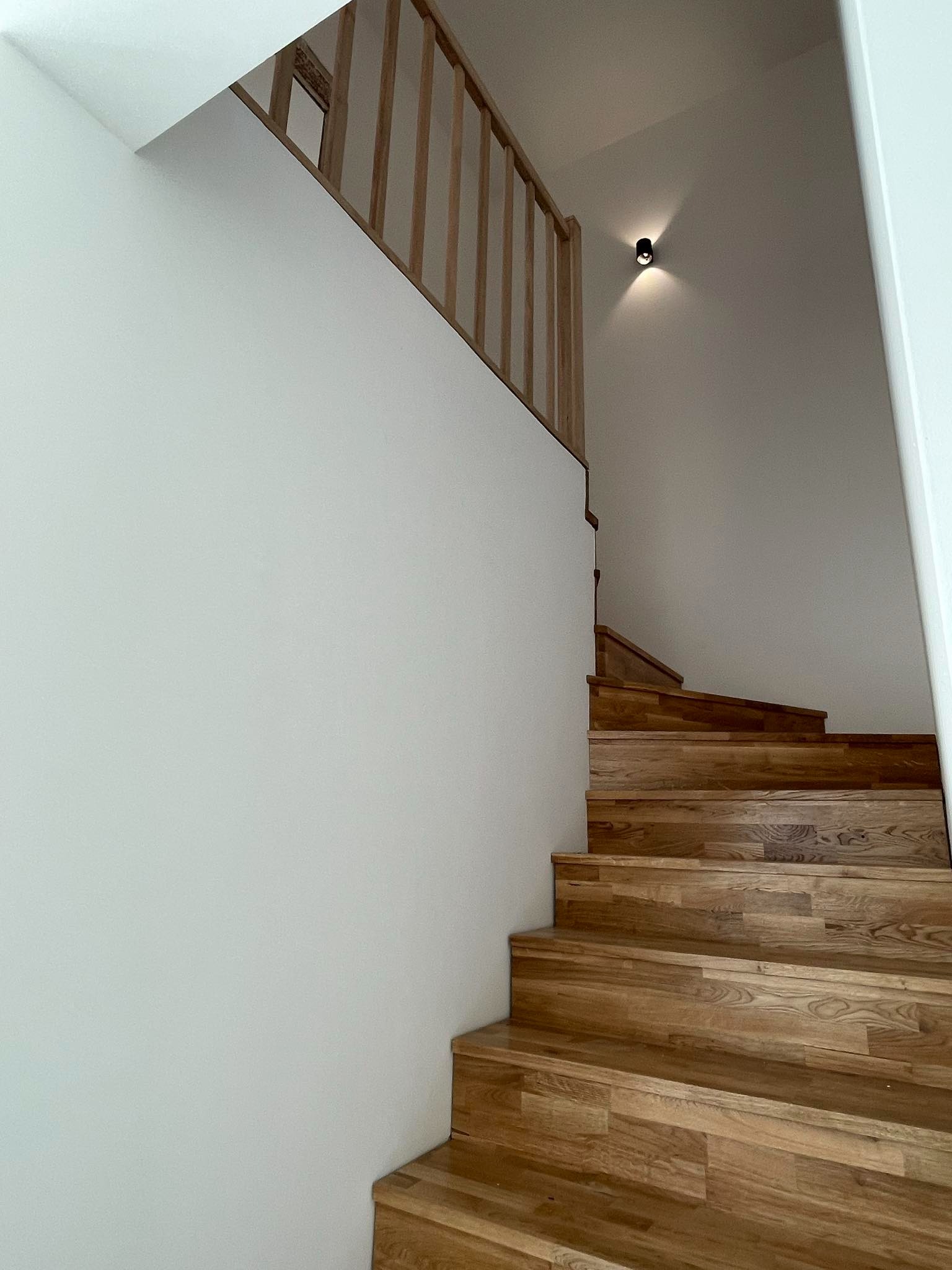 Schody drewniane z jasnymi stopniami i balustradą, prowadzące w górę przy białej ścianie, oświetlone punktowym światłem.