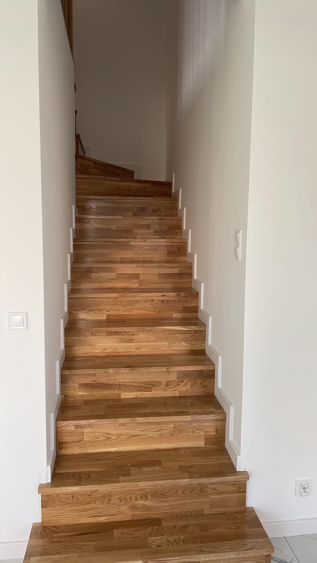 Drewniane schody z białymi listwami przypodłogowymi, prowadzące na górę, z widocznymi włącznikami światła na białej ścianie.