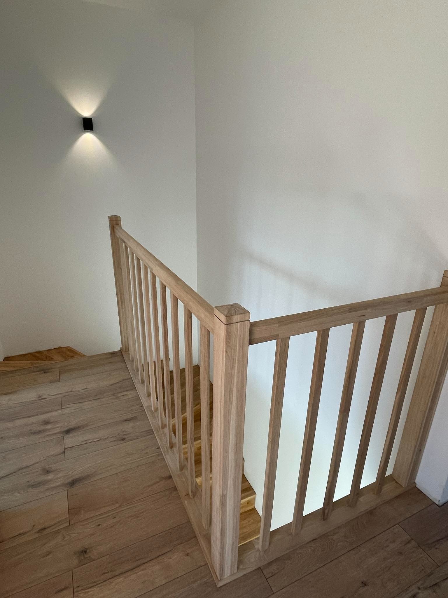 Drewniane schody z jasną balustradą, oświetlone kinkietem na białej ścianie, widok z góry na parkiet.